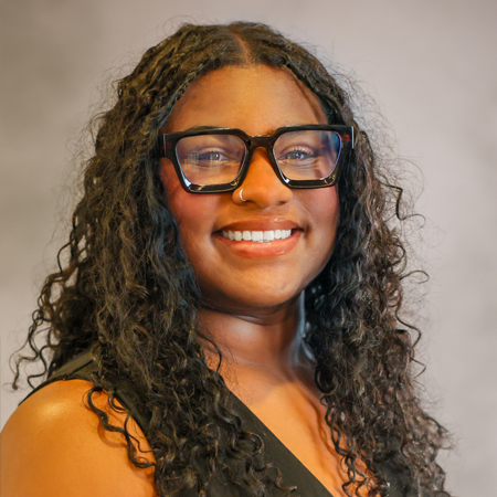 Professional headshot of a smiling person with long, curly black hair wearing oversized black rectangular eyeglasses and a sleeveless black blouse. The person is facing the camera and is wearing a small nose ring. The photo is set against a soft gray studio background with warm, even lighting, creating a friendly, approachable, and professional appearance.