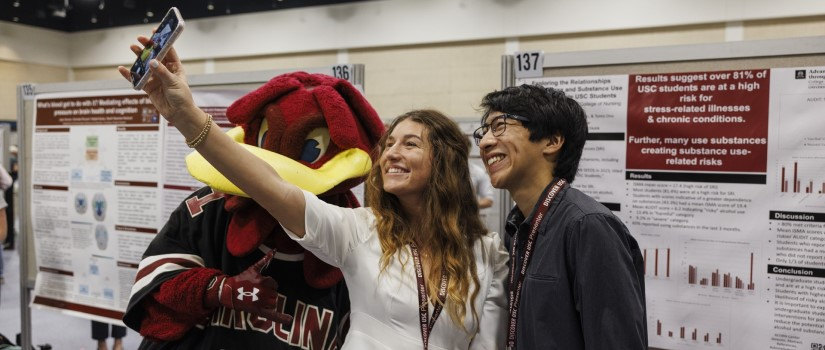 At a Discover USC poster session, Cocky—the University of South Carolina’s Gamecock mascot—stands taking a selfie with two smiling Discover USC presenters.