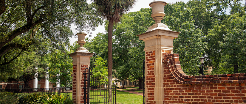 A person rides a bicycle along a shaded brick walkway on the University of South Carolina Horseshoe, surrounded by tall green trees and campus lampposts. A banner on one lamppost reads “47 nationally ranked programs.