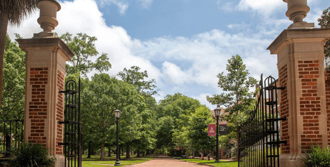 View through the historic iron gates of the Horseshoe at the University of South Carolina, showing a tree-lined brick path with classic lampposts and a banner displaying the university logo.