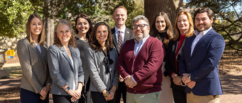 A group of nine professionally dressed individuals posing outdoors on a sunny day, smiling at the camera. They are standing in front of a large tree with a background of greenery and a park-like setting.