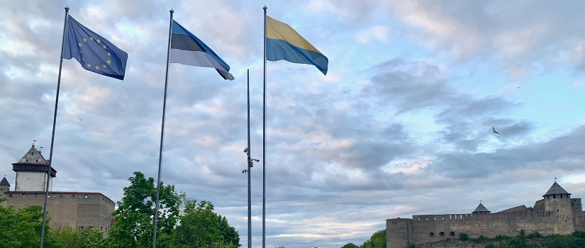 Three flags — the NATO, Estonian, and Narva City flag - fly next to the Narva River. Hermann Castle stands in Estonia on the left side of the photo and across the Narva River in Russia there is the Ivangorod Fortress. Photo by Kara Brown, June 2025.