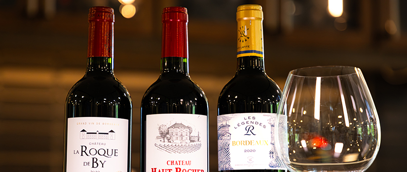 Three bottles of Bordeaux wine displayed beside an empty wine glass on a wooden table, with a softly lit, blurred background creating a warm, elegant tasting room atmosphere.