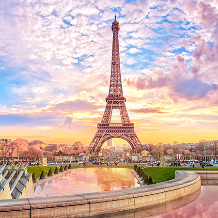 The Eiffel Tower rises above fountains and gardens at sunset in Paris, with pink and golden hues filling the sky.