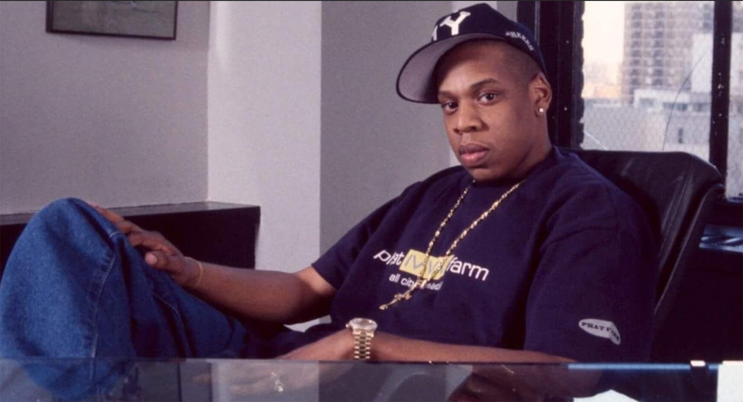 A seated African American adult male in black t-shirt and ballcap looks into the camera