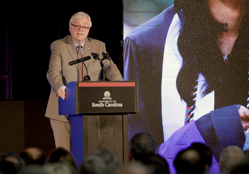 a man speaks at a podium with images on a screen in the background