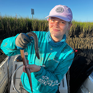 Young woman in blue shirt and ponytail holding a snake in a coastal marsh setting